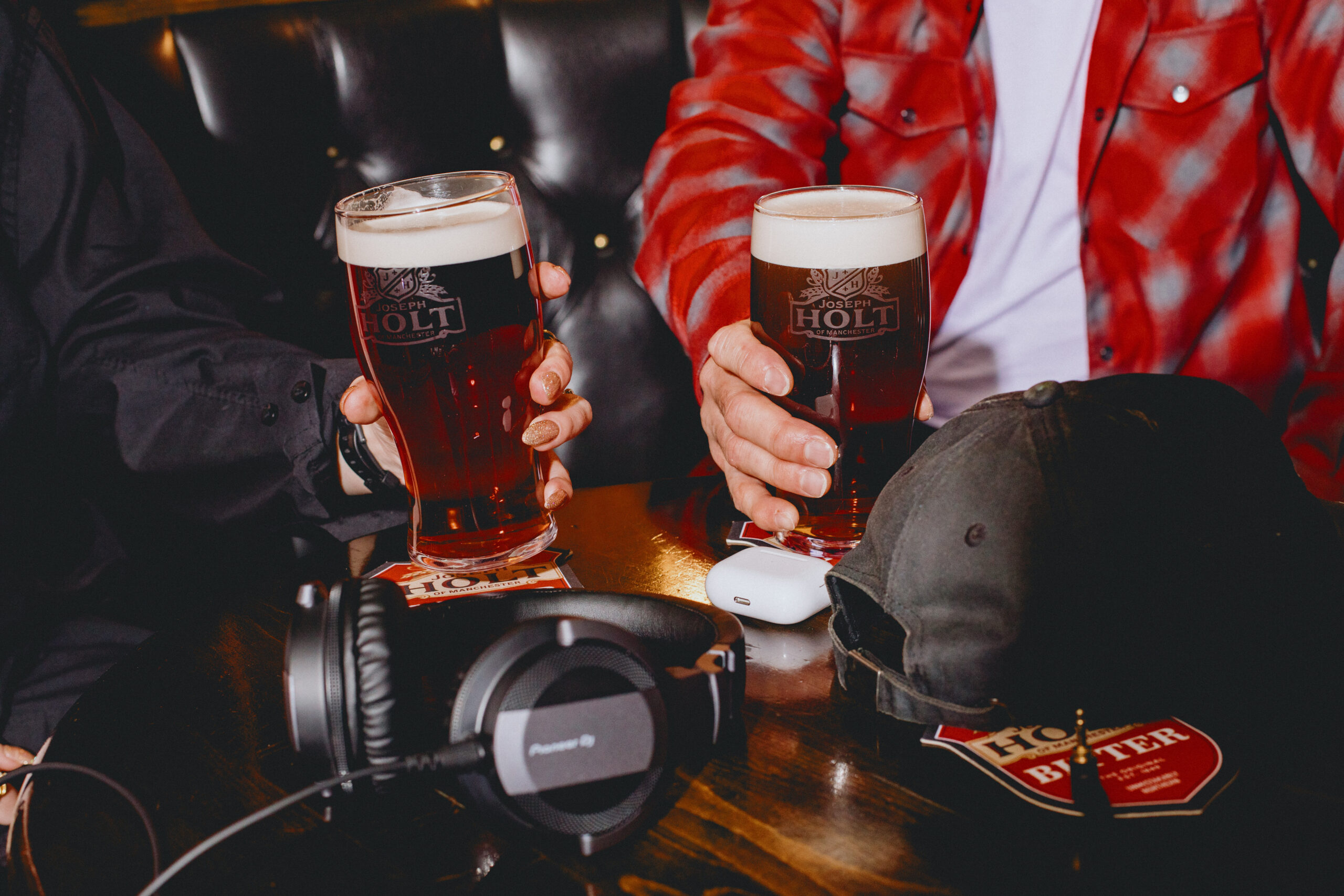 Two pints of bitter next to some headphones, airpods and a hat